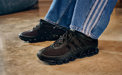 The image shows a pair of dark-colored hiking boots or sneakers worn by a person, with the person's jeans-clad legs visible in the foreground. The background appears to be a wooden or tiled floor.