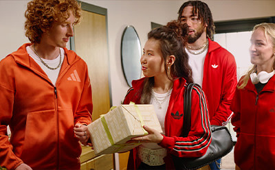 The image shows a group of young people, two men and two women, standing together in what appears to be a hallway or entryway. They are all wearing red and orange clothing, and one of the women is holding a book or folder.