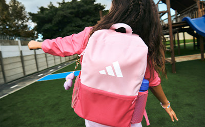 A young person wearing a pink and red backpack with the Adidas logo, standing in front of a grassy field and fenced area.
