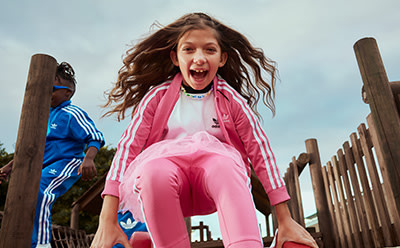 A young woman with long, curly hair is enthusiastically jumping and smiling in a pink outfit, with a wooden fence and trees in the background.