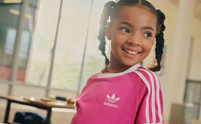 A young girl with braided hair wearing a pink Adidas shirt is smiling brightly in what appears to be an indoor setting with a window in the background.