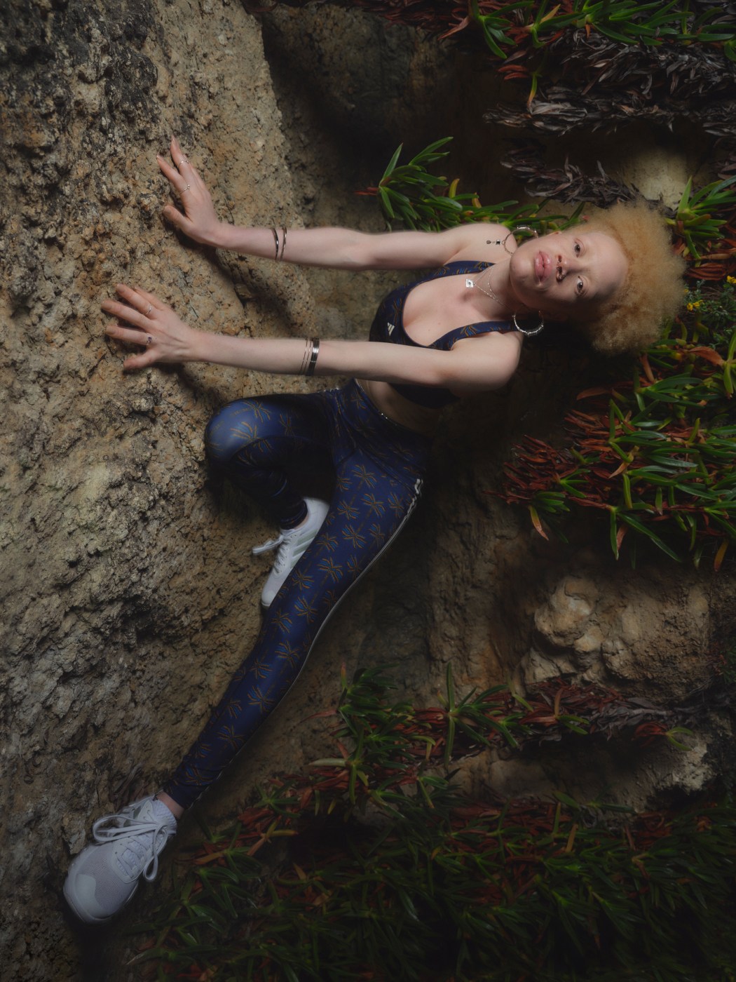 Model wearing adidas X FARM Rio navy blue printed sports bra and matching leggings. She is perched on a sandy beach rock.