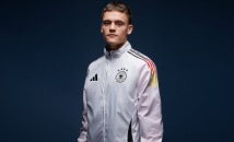 Mladý muž v bielej športovej bunde s logom Nemeckého národného futbalového mužstva stojí na tmavom pozadí.