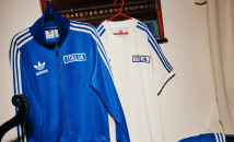 De afbeelding toont twee adidas sportshirts, een in blauw en een in wit, die aan een kledingrek hangen tegen een houten achtergrond.