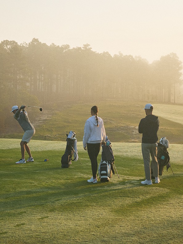 Skupina golfistov sa hrá na bujnom zelenom golfovom ihrisku obklopenom hmlou zalesnenou krajinou v pozadí.