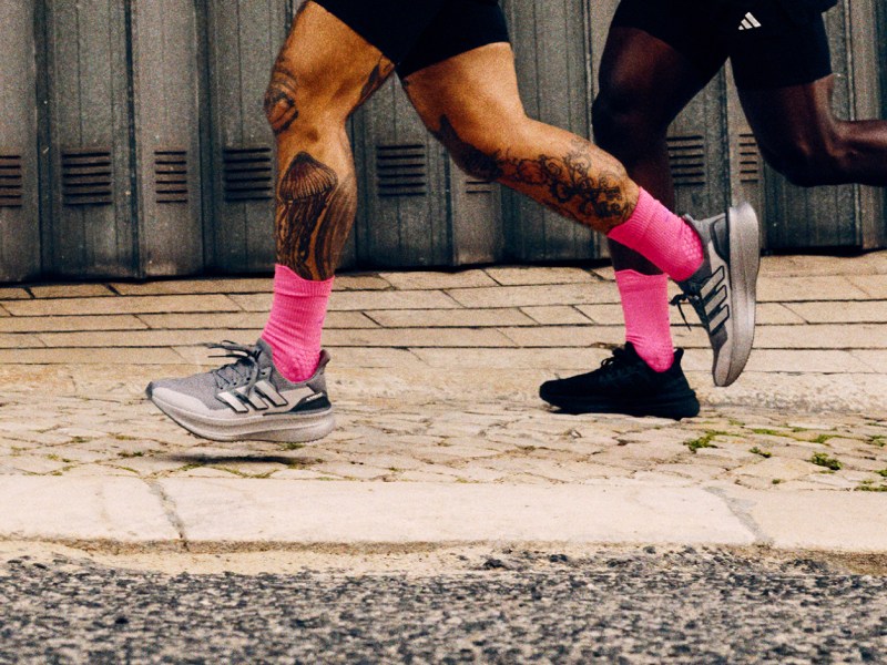 The image shows a person's legs and feet wearing athletic shoes and bright pink socks, standing on a paved surface with a brick wall in the background.