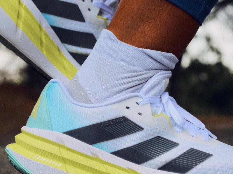 The image shows a close-up view of a person's feet wearing a pair of white and teal athletic shoes with yellow accents, standing on a blurred background.