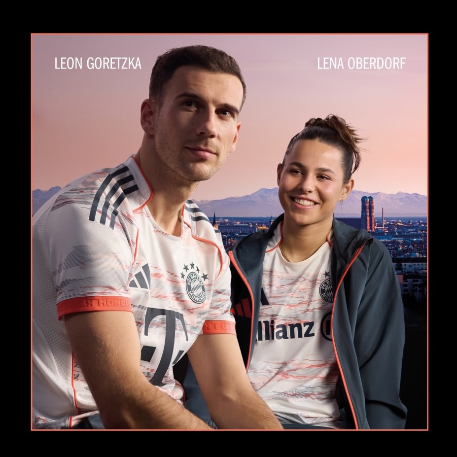 The image shows two individuals, a man and a woman, posing together in front of a cityscape backdrop. The man is wearing a white and red sports jersey, while the woman is wearing a gray jacket. The background features a skyline with tall buildings and a colorful sky.
