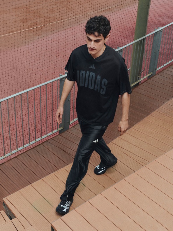 A young man wearing a black t-shirt and jeans is standing on a wooden deck, with a metal railing visible in the background.