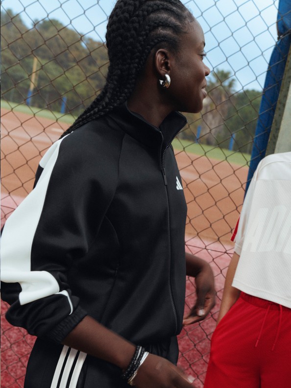 A person wearing a black and white athletic jacket stands in front of a chain-link fence, with a sports field visible in the background.