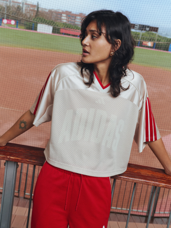 A young woman with dark curly hair wearing a white and red sports jersey stands on a baseball field with a stadium in the background.