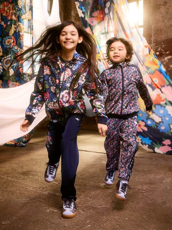 Two young girls wearing colorful, floral-patterned outfits are walking together in a vibrant, artistic environment with murals and other decorative elements in the background.