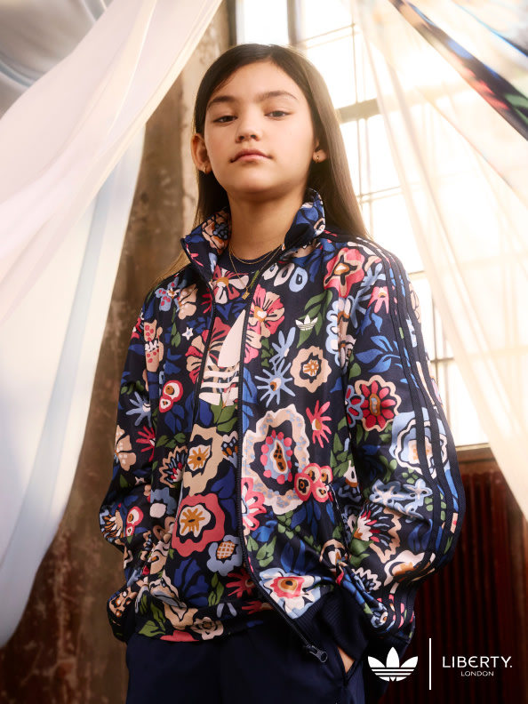 A young woman wearing a colorful floral-patterned jacket stands in a room with a white curtain backdrop.