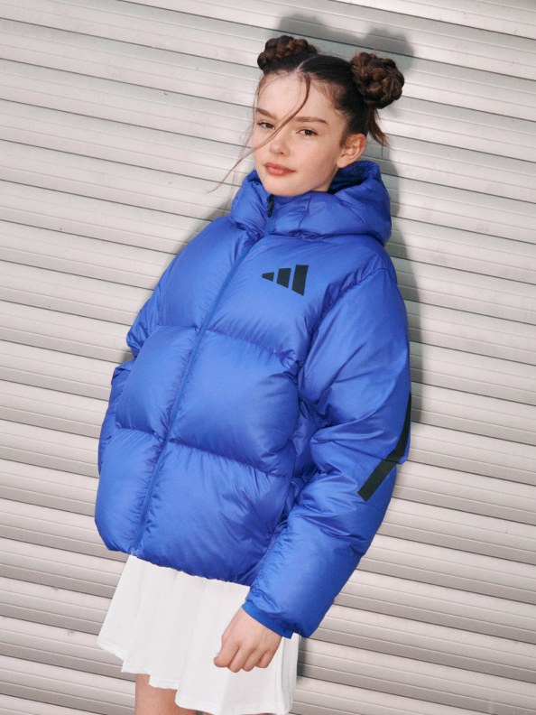 A person wearing a bright blue puffy jacket stands in front of a white corrugated metal wall.