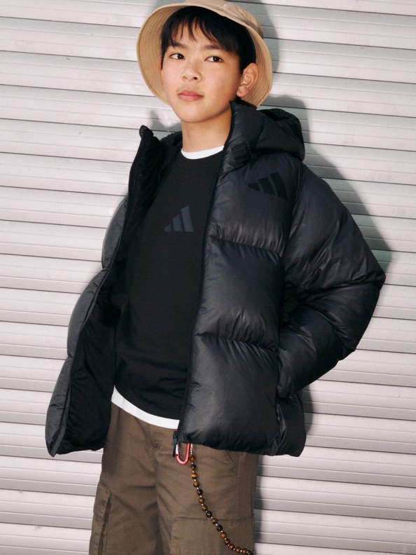 A young person wearing a black puffy jacket and a beige hat stands in front of a white wall.