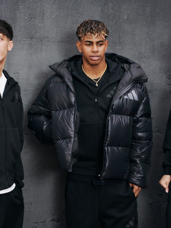 The image shows a young man wearing a black puffy jacket standing in front of a gray concrete wall.