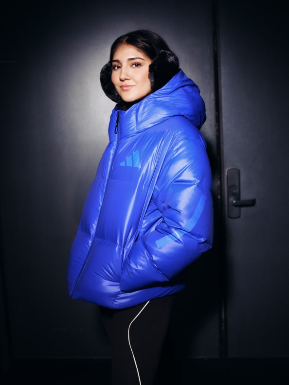 A person wearing a bright blue puffy jacket stands in a dimly lit doorway, their face partially obscured by the jacket's hood.
