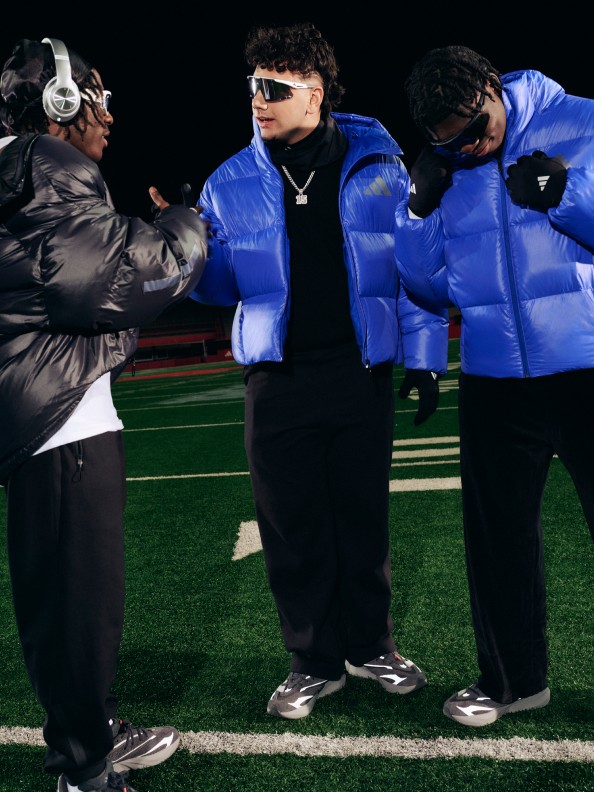 青いダウンジャケットとサングラスを身に着けた3人が、草の生えた野原に立ち、背後にダークジャケットを着けた人物が立っている。