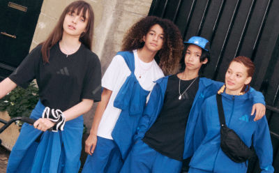 The image shows a group of four young women standing together, wearing a mix of casual and athletic clothing, with one woman wearing a blue jacket and the others in darker colors. They appear to be standing in front of a dark background, possibly a wall or building.