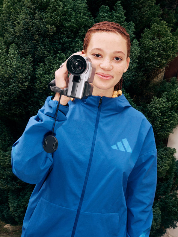 A young person wearing a blue hooded jacket is holding a video camera and smiling while standing in a forested area.