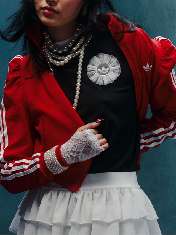 A person wearing a red jacket with white stripes, a black top, and a pearl necklace stands against a teal background.