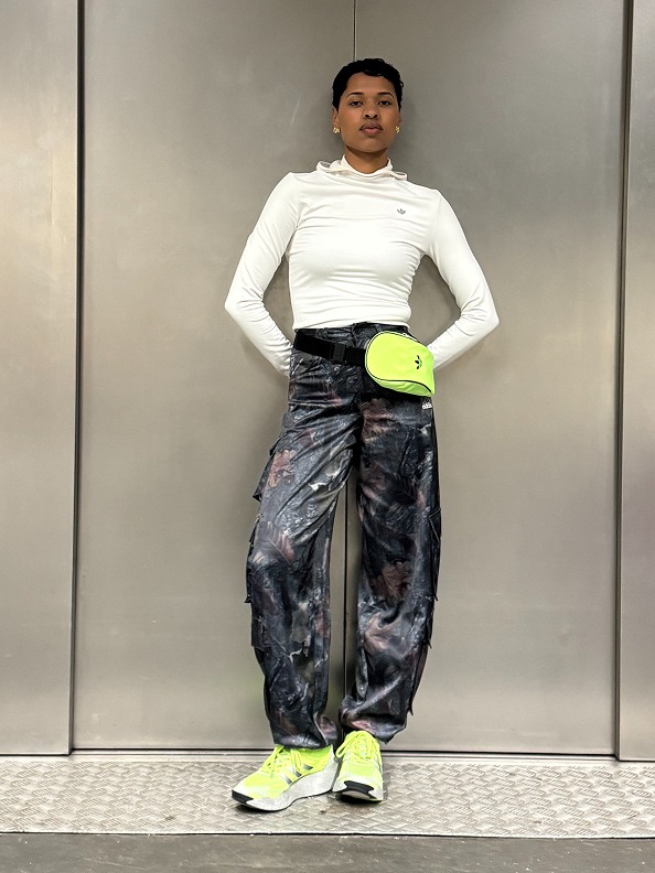 A person wearing a white long-sleeved top, dark-patterned pants, and bright yellow sneakers stands in front of a gray wall.