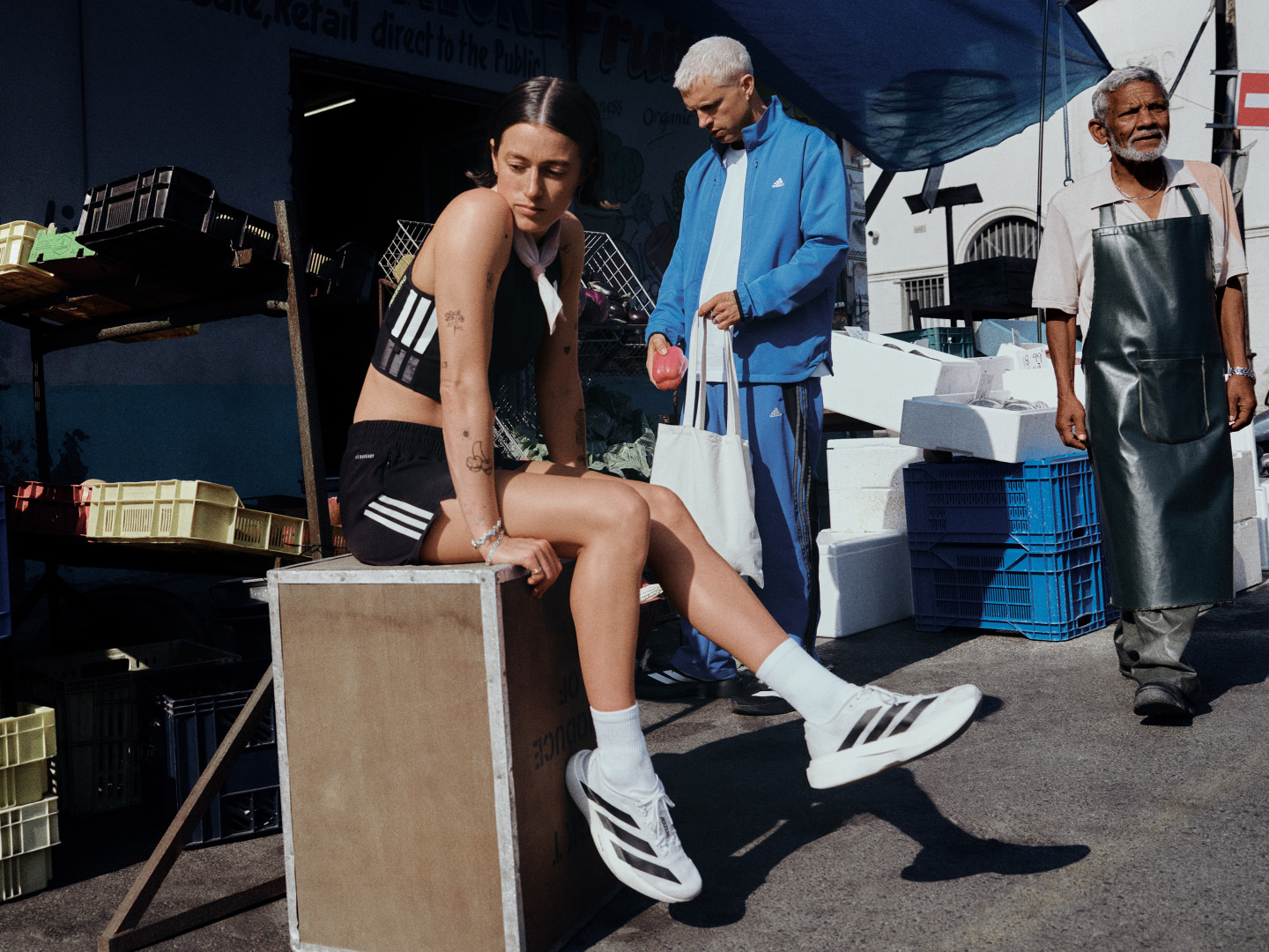 The image depicts a person sitting on a crate, wearing athletic clothing, surrounded by various objects and individuals in what appears to be a backstage or behind-the-scenes setting.
