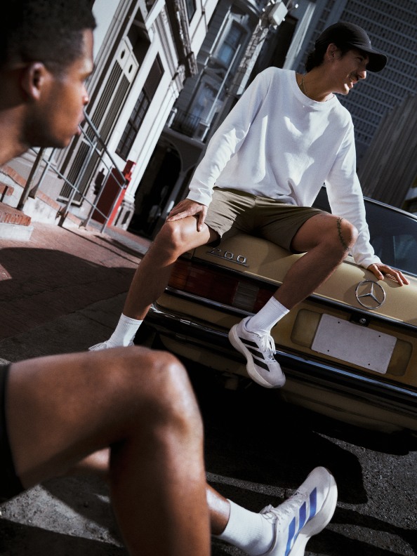 Two individuals, one seated and one standing, are engaged in an interaction in an urban setting with buildings and structures visible in the background.
