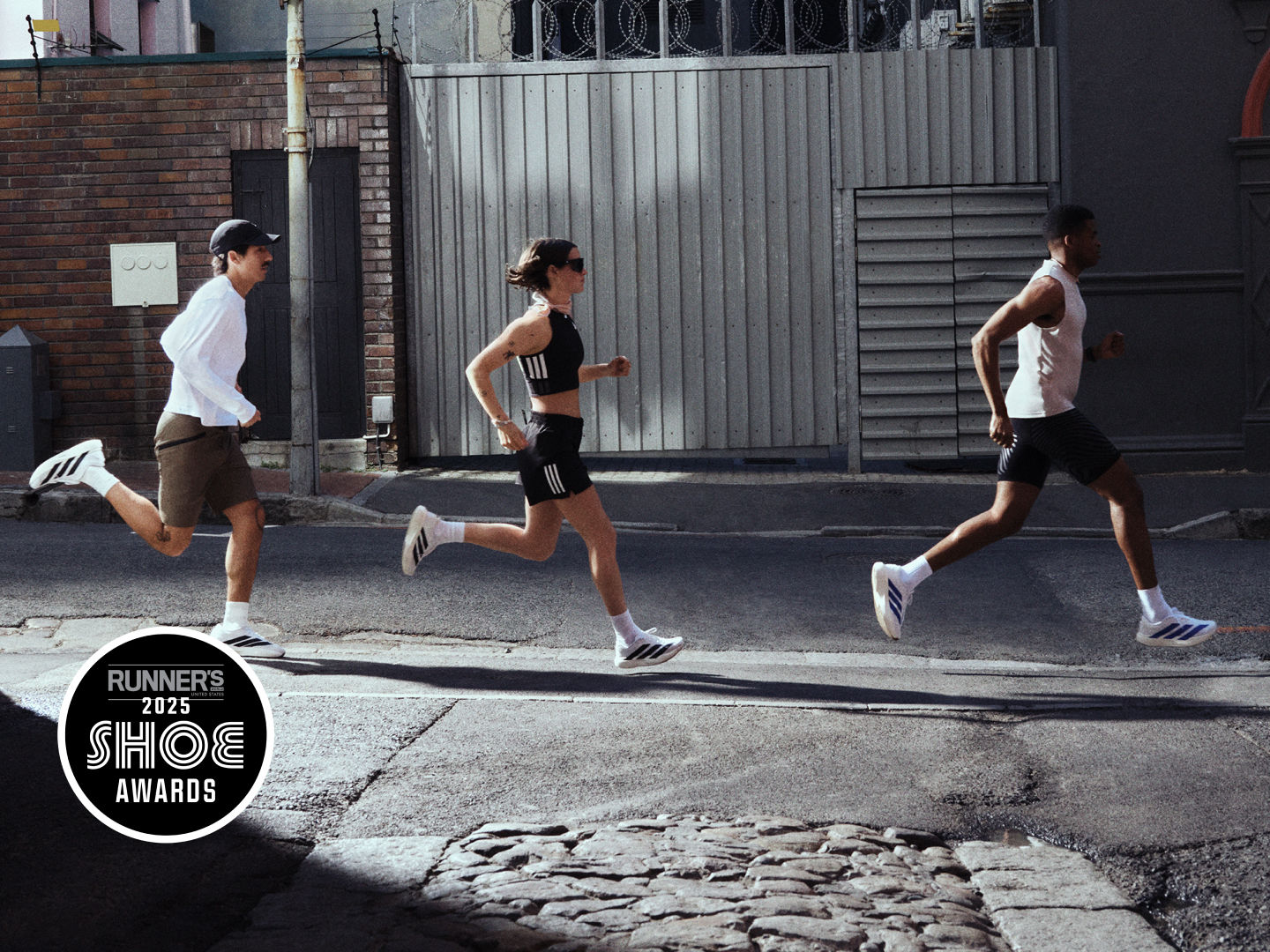 Three runners in athletic gear are jogging on a city street, with a brick building and a metal gate visible in the background.