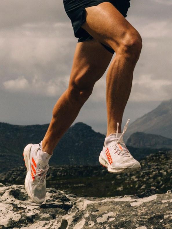 Muskularne nogi w sportowych butach stoją na skalistym, górskim terenie na pochmurnym niebie.