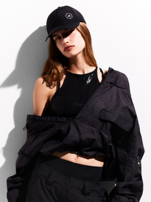 A young woman wearing a black baseball cap and a black crop top stands against a plain white background, her expression serious and her gaze directed away from the camera.