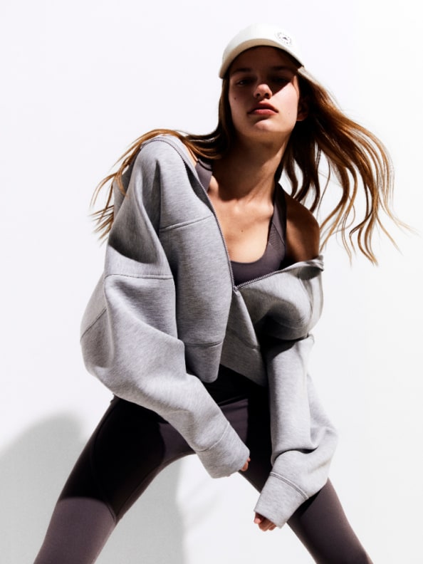 A young woman with long, windblown hair wearing a gray sweatshirt and black leggings stands against a plain white background.
