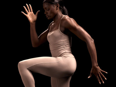 Una figura femenina con un body beige es capturada en una pose dramática sobre un fondo oscuro, sus brazos levantados y su cuerpo en movimiento.