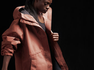 Una persona con una chaqueta con capucha rosa está de pie sobre un fondo oscuro, con la cara parcialmente oscurecida por la capucha.