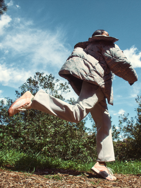 Eine Person mit breitem Schirm und lässiger Kleidung läuft durch ein Grasfeld mit Bäumen im Hintergrund vor einem blauen Himmel mit Wolken.