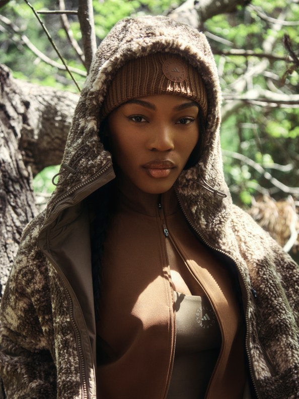 Eine Person mit einer Kapuzenjacke steht in einem Waldgebiet, im Hintergrund sind Bäume und Äste zu sehen.