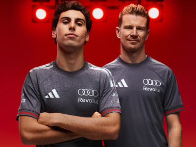 Two individuals wearing gray sports jerseys with the Audi logo, standing in front of a red background with bright lights.