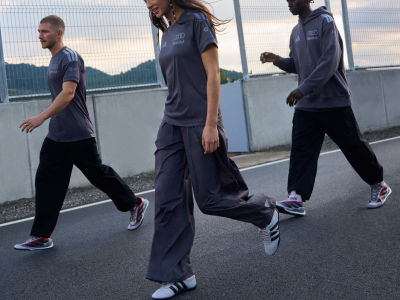 Three individuals wearing dark-colored athletic clothing and sneakers are walking on a paved road, with a mountainous landscape visible in the background.