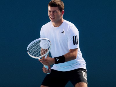 Ein junger Mann in einem weißen sportlichen Hemd und schwarzen Shorts hält einen Tennisschläger und scheint sich mitten in einem Tennisspiel mit dunkelblauem Hintergrund zu befinden.