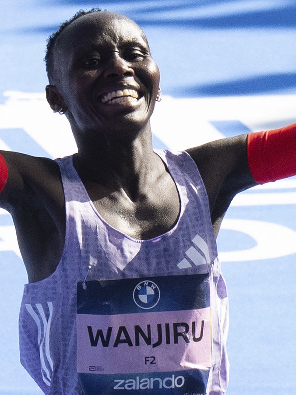 A smiling Black athlete wearing a running jersey with the name "Wanjiru" on it, against a bright blue sky background.