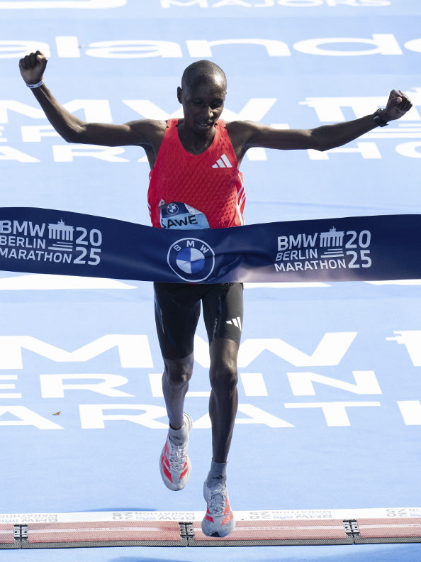 A male runner in a red jersey crosses the finish line of the BMW Berlin Marathon, with his arms raised in celebration against a backdrop of the event's branding.