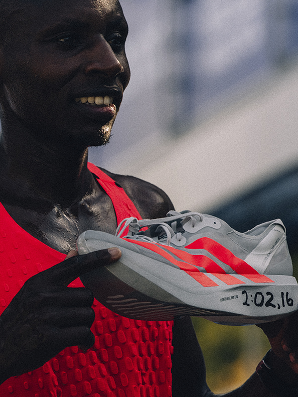 A close-up portrait of a smiling Black man wearing a red shirt and holding a pair of running shoes with the date "2:02.16" written on them.