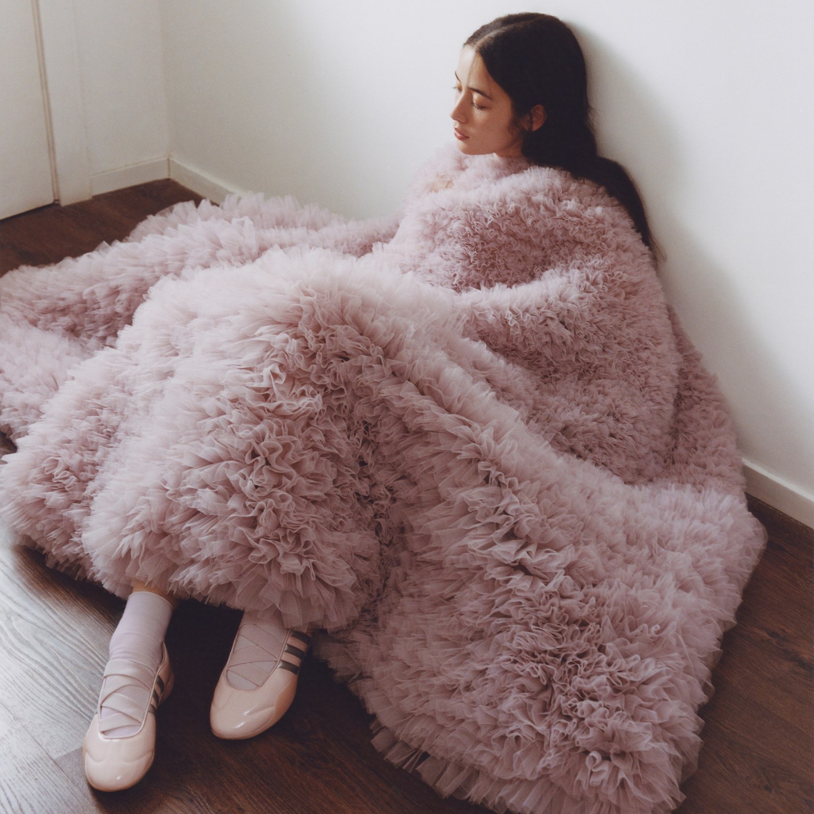 ผู้หญิงคนหนึ่งนั่งอยู่บนพื้นไม้ห่อด้วยผ้าห่มสีชมพูนุ่มฟู เธอสวมรองเท้าเทควันโด CLOT สีชมพูอ่อนจากอาดิดาส แต่งแถบสีเงินเมทัลลิกที่ออกแบบโดย Caroline Hu