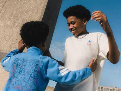 Two young individuals, one wearing a blue hoodie and the other a white t-shirt, are interacting with each other in an outdoor setting with a concrete wall and a clear blue sky in the background.