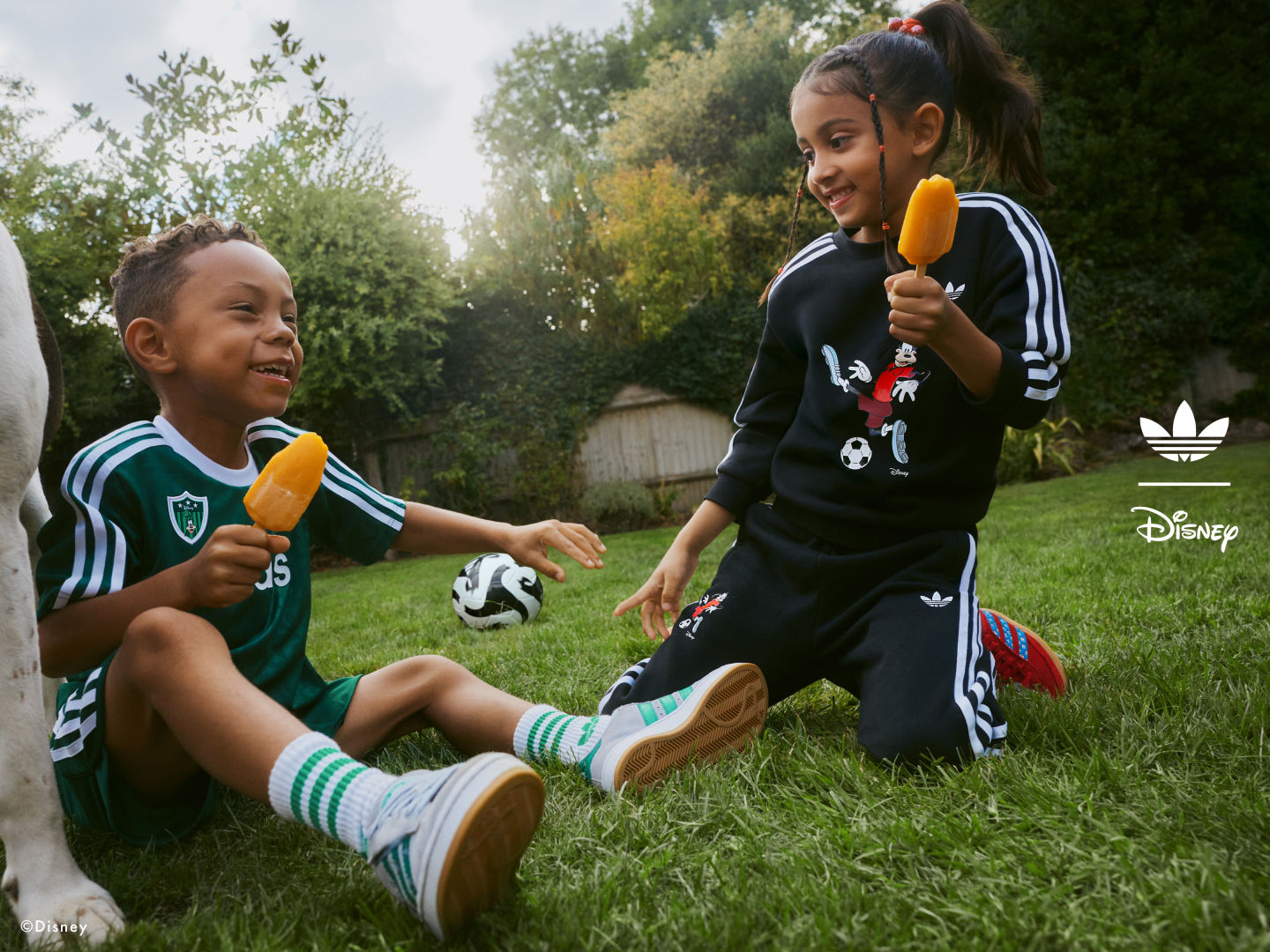 Two young children, a boy and a girl, are playing with soccer balls on a grassy field surrounded by trees and foliage.
