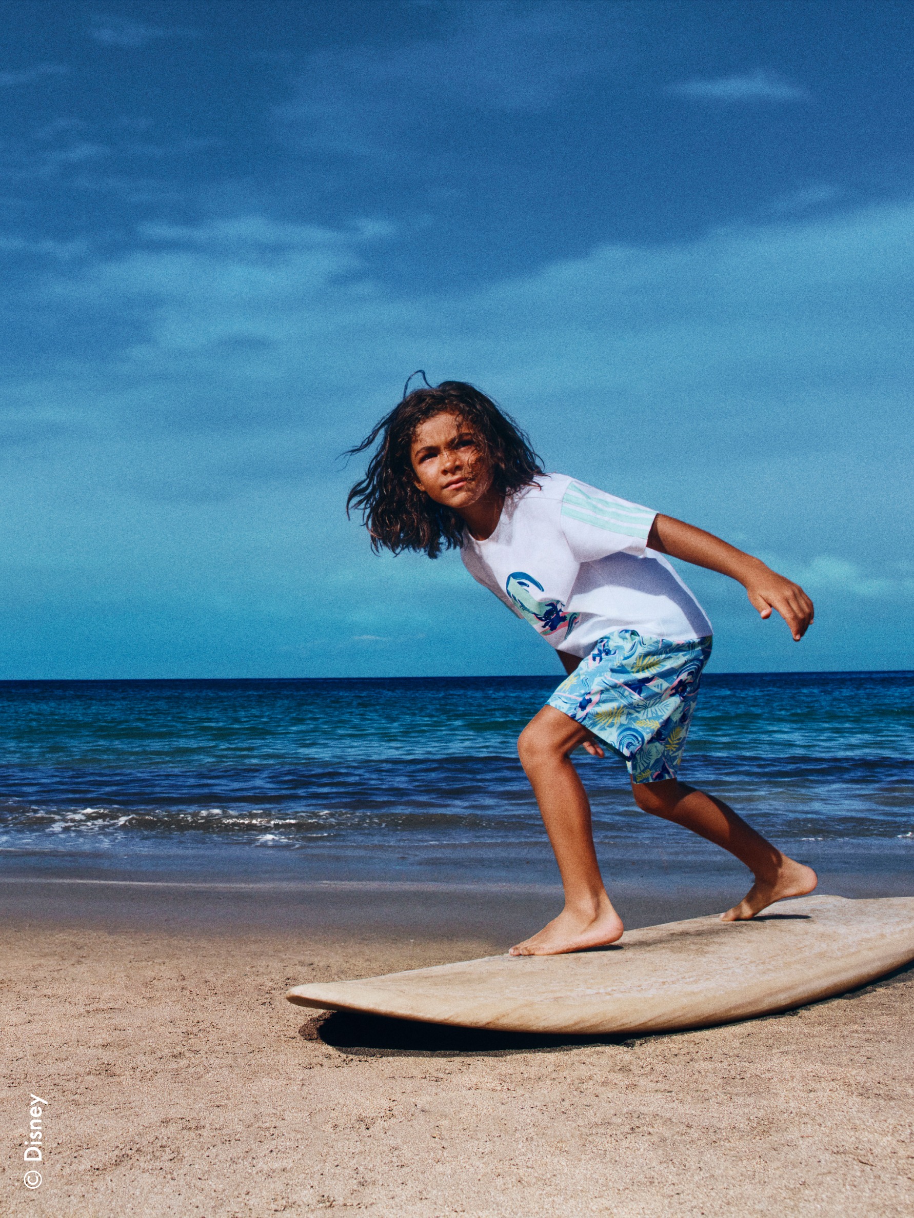 A young person in casual beachwear is riding a surfboard on a sandy beach with a vast, blue ocean and clear sky in the background.