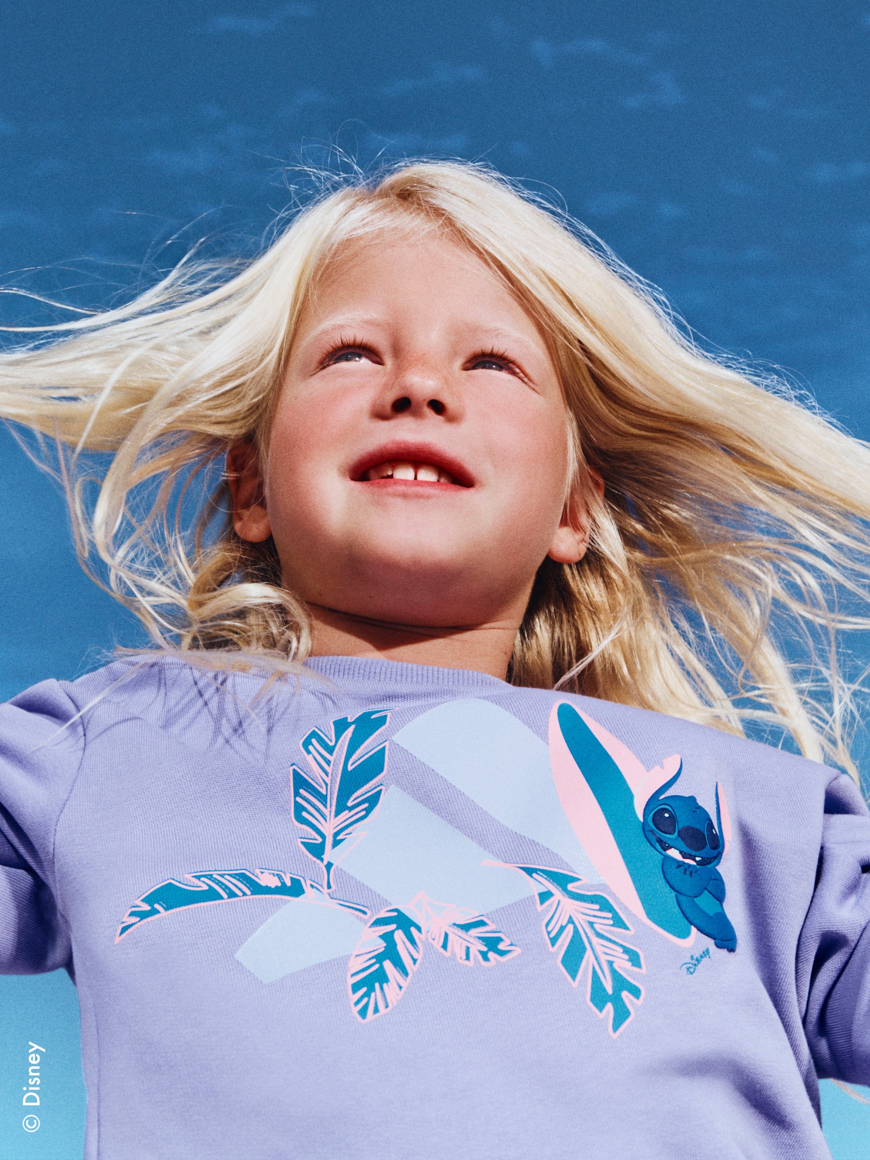 A young girl with flowing blonde hair smiles joyfully against a vibrant blue sky, wearing a shirt with a whimsical feather and Stitch character design.