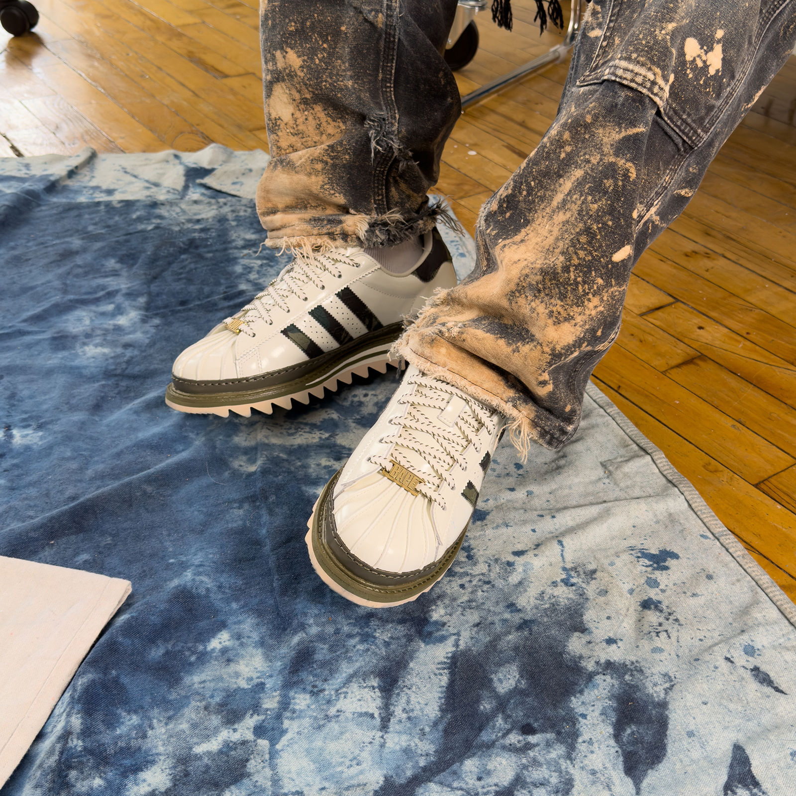 A pair of adidas CLOT x BAPE Superstar sneakers on a light wooden surface with blue paint marks. One shoe shows the black star logo, and the other displays the adidas label and Three Stripes.