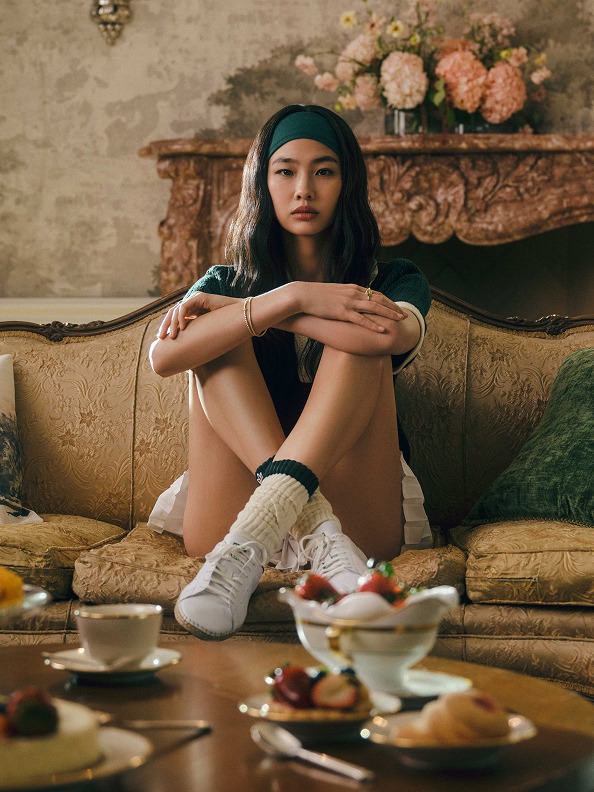 A woman sits cross-legged on a sofa wearing white adidas x Edison Chen CLOT Stan Smith sneakers, a green and white polo shirt, long white socks with green trim, and a green headband from the adidas x Edison Chen CLOT Stan Smith collection.