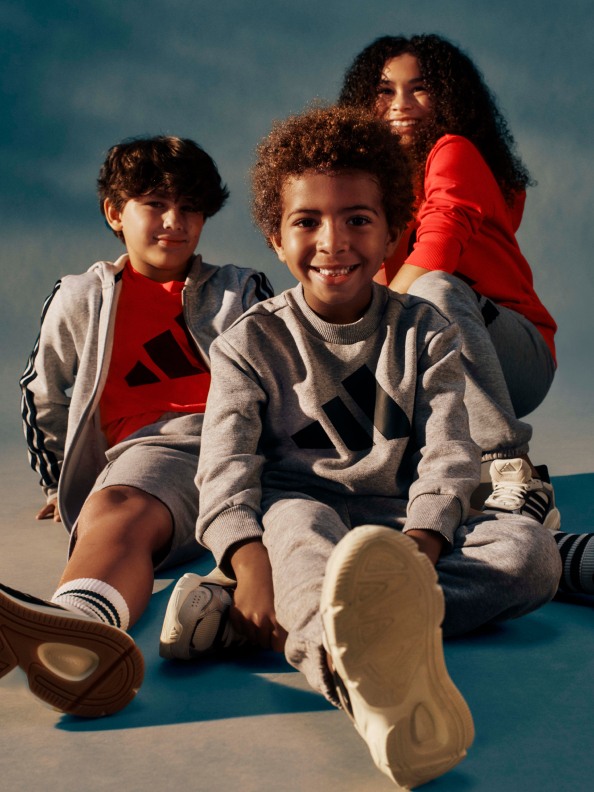 Three children, two boys and a girl, sit together on a colorful surface, smiling and appearing to be enjoying themselves in an outdoor setting with a cloudy sky in the background.